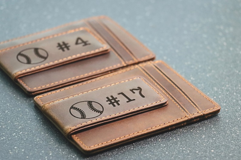 Puede incluir: Dos carteras de cuero marr&oacute;n con un b&eacute;isbol y un n&uacute;mero grabados en la parte delantera. La cartera superior tiene el n&uacute;mero "#4" y la cartera inferior tiene el n&uacute;mero "#17".