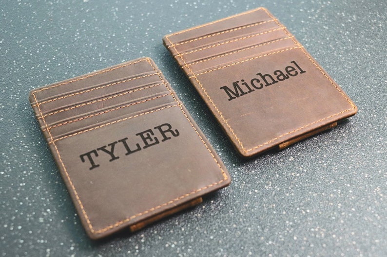 Puede incluir: Dos carteras de cuero marr&oacute;n con un clip para billetes. La cartera de la izquierda tiene el nombre "TYLER" grabado en relieve. La cartera de la derecha tiene el nombre "Michael" grabado en relieve.