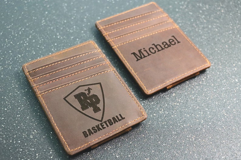 Puede incluir: Dos carteras de cuero marr&oacute;n con un clip para billetes. Una cartera tiene el texto "Michael" grabado en ella. La otra cartera tiene un escudo con las letras "RP" y el texto "BASKETBALL" grabado en ella.