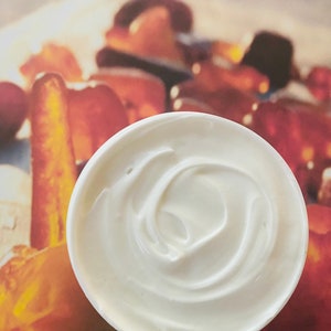 May include: A white plastic cup filled with a swirl of white creamy sauce, likely sour cream or mayonnaise, on a background of orange and brown.