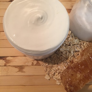 May include: A white jar of body butter with a swirl on top, next to a pile of oats and a piece of honeycomb.