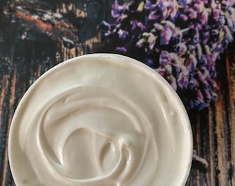 Może przedstawiać: Biały, kremowy produkt do pielęgnacji skóry w okrągłym pojemniku. Produkt ma wirującą konsystencję. Gałązka fioletowej lawendy znajduje się w tle, na tle ciemnej powierzchni z usłojeniem drewna.