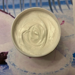 May include: A white, creamy, whipped body butter in a white plastic container. The butter is swirled and has a smooth, even texture.