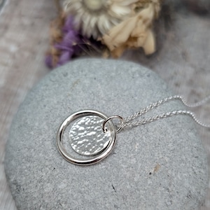 Peut inclure: Un collier en argent avec un pendentif rond martelé et une petite bague circulaire autour. Le collier est posé sur une surface texturée grise.