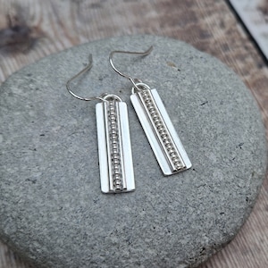 Puede incluir: Un par de pendientes colgantes de plata con un diseño de cuentas. Los pendientes son de forma rectangular y tienen un estilo simple y moderno.