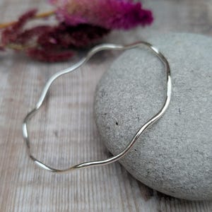 May include: A silver bangle bracelet with a wavy design. The bracelet is lying on a grey stone.