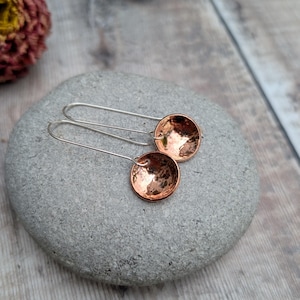 May include: Handmade copper earrings with a hammered finish. The earrings have a circular, bowl-like shape, suspended from long, silver-coloured ear wires. The copper has a warm, reddish-brown colour, and the earrings are displayed on a grey stone.