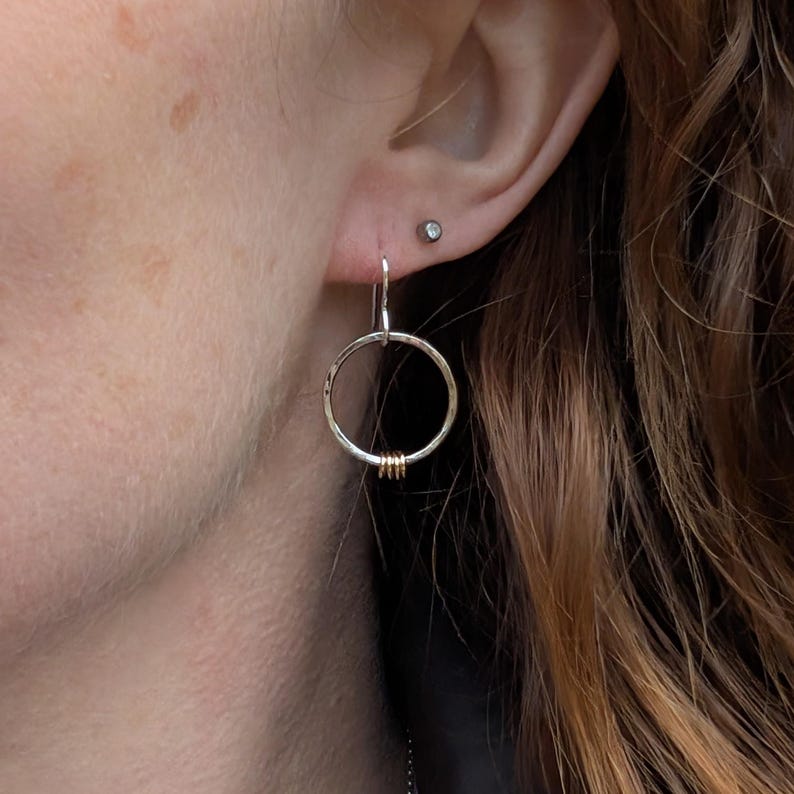 One silver and gold-accented circle earring being worn. The hammered silver hoop dangles just below the earlobe, with the small gold loops adding a warm contrast.