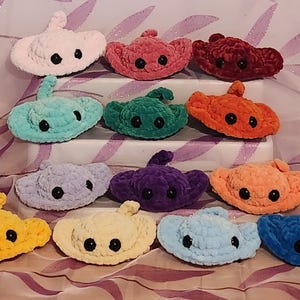 May include: A collection of 12 crocheted stingray plushies in various colors, including pink, red, green, orange, purple, blue, and yellow. Each stingray has black eyes and a crocheted texture.