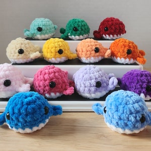 May include: A collection of fourteen small, plush, crocheted whale toys in various colors including light blue, green, red, cream, yellow, peach, orange, pink, magenta, lavender, purple, and dark blue, each featuring black plastic safety eyes.