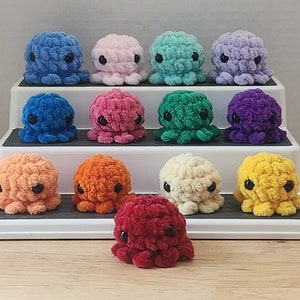 May include: A set of 12 crocheted octopus plushies in various colors, displayed on a white tiered shelf. The octopus plushies are made with yarn in shades of blue, pink, green, purple, orange, yellow, and red.