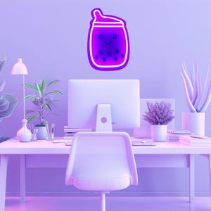 May include: A purple neon sign shaped like a boba tea cup with a cute face and a straw. The sign is hanging on a wall in a home office setting with a computer, desk, and plants.