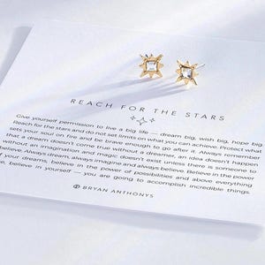 Puede incluir: Pendientes de estrella dorados con cristales transparentes. Los pendientes están sobre una tarjeta blanca con el texto "REACH FOR THE STARS" y una cita sobre soñar a lo grande.