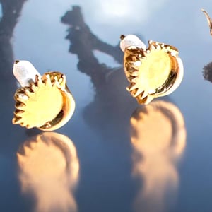 May include: A pair of gold-colored stud earrings. Each earring features a sunburst design with a yellow center and a white post. The earrings are displayed on a reflective surface, creating a mirrored effect.