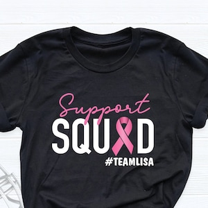 May include: Black t-shirt with white text that reads "Support Squad" with a pink ribbon and the hashtag "#TeamLisa".