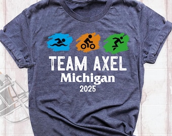 May include: A dark blue t-shirt with a white design. The design includes three icons representing swimming, biking, and running. The text "TEAM AXEL Michigan 2025" is printed below the icons.
