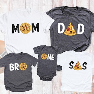 May include: A set of four t-shirts with a pizza theme. The shirts say "MOM", "DAD", "BRO", and "SAS" with a pizza slice graphic next to each word. The shirts are in white and gray.