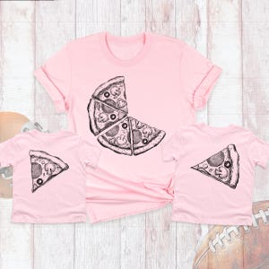 May include: Two pink t-shirts with a pizza slice graphic. The larger shirt has a graphic of three pizza slices, while the smaller shirt has a graphic of one pizza slice.