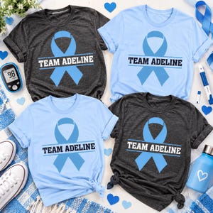 May include: Four short-sleeved t-shirts in charcoal grey and light blue, each featuring a light blue awareness ribbon graphic and the text "TEAM ADELINE" printed in bold, dark lettering across the centre of the chest.
