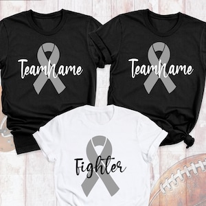 May include: Three t-shirts: two black and one white. Each shirt features a gray awareness ribbon. The black shirts have the text "Team Name" in white script. The white shirt says "Fighter" in black script.