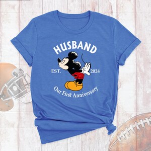 Puede incluir: Una camiseta azul con un gr&aacute;fico blanco de Mickey Mouse dando un beso. El gr&aacute;fico tambi&eacute;n incluye el texto "Husband Est. 2024 Our First Anniversary".
