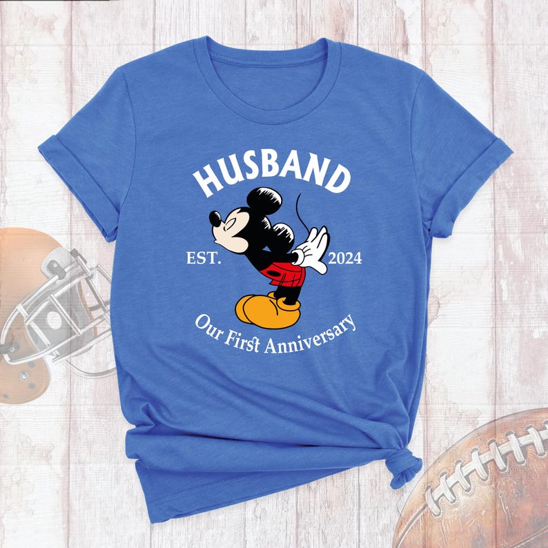Puede incluir: Una camiseta azul con un gr&aacute;fico blanco de Mickey Mouse dando un beso. El gr&aacute;fico tambi&eacute;n incluye el texto "Husband Est. 2024 Our First Anniversary".