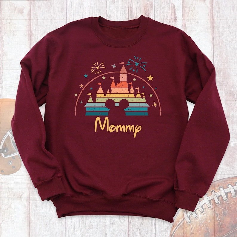 May include: Burgundy sweatshirt with a retro-style rainbow castle graphic and the word "Mommy" underneath. The castle has a Mickey Mouse silhouette in the center.