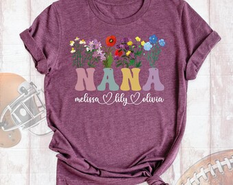 May include: A heathered maroon short-sleeve t-shirt featuring a graphic of colorful wildflowers emerging from the letters of the word NANA. Below the word, the names Melissa, Lily, and Olivia are printed in white script with heart icons.