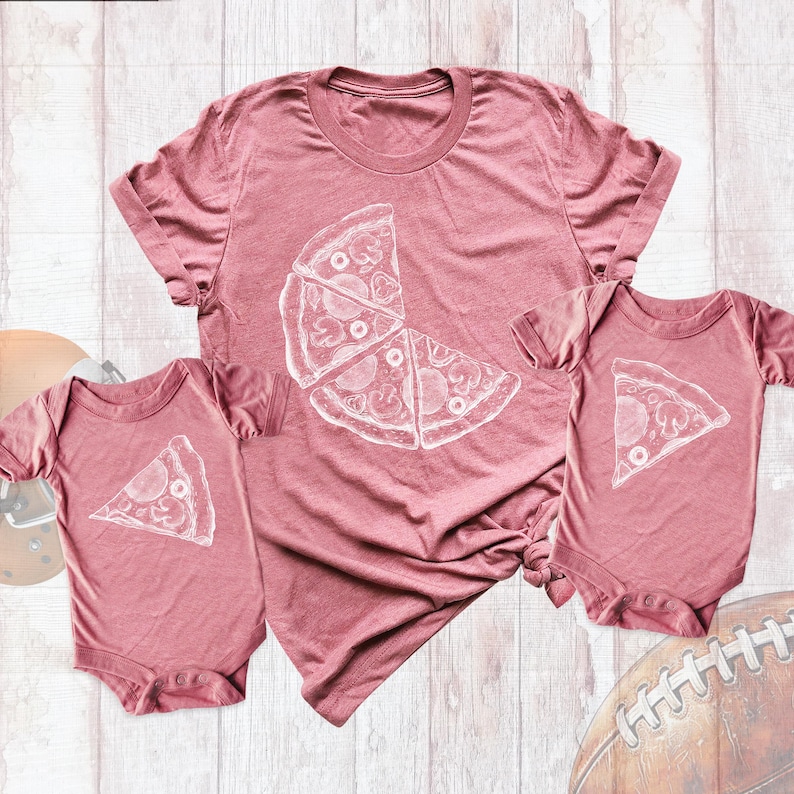 May include: Two pink T-shirts with a white pizza graphic. The larger shirt has a half pizza with three slices, and the smaller shirt has a single slice of pizza.