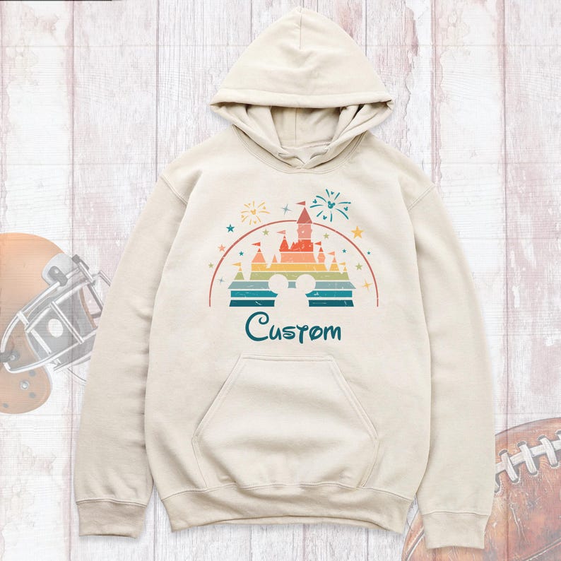 May include: A beige hoodie with a vintage-style graphic of a castle with fireworks and the word "Custom" printed below.