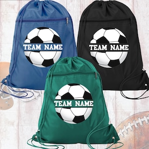 May include: Three drawstring sports bags in blue, black, and green. Each bag features a soccer ball graphic with the text "TEAM NAME" in white. The bags have a zippered pocket and a drawstring closure.