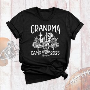 May include: Black t-shirt with white text that reads "GRANDMA CAMP 2025" above a graphic of two Adirondack chairs facing a campfire, with trees in the background. The name "Emilia" is written below the campfire.