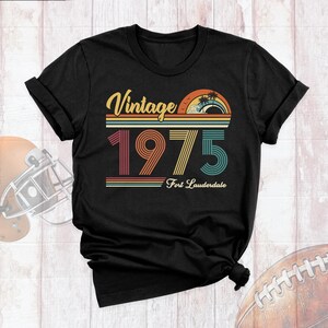 May include: Black t-shirt with a vintage-style graphic that says "Vintage 1975 Fort Lauderdale". The graphic features a sunset with palm trees and a wave.