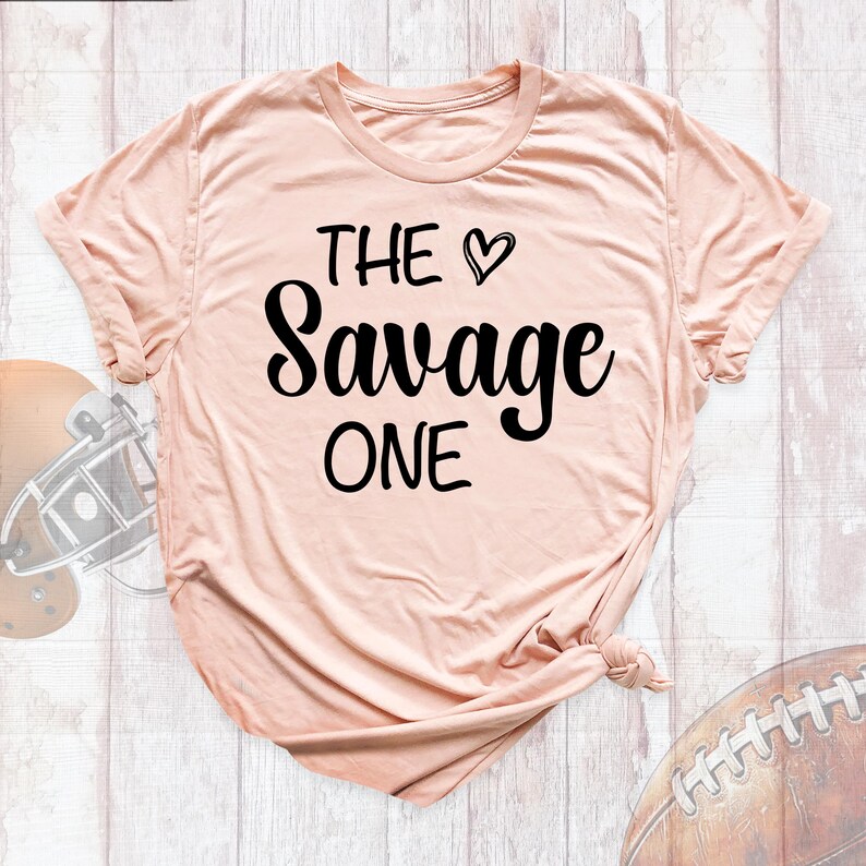 May include: A peach-colored t-shirt with the text "THE &hearts; Savage ONE" printed in black.