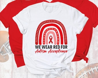 May include: A white short-sleeved t-shirt featuring a red rainbow graphic with a red awareness ribbon in the center. The text reads WE WEAR RED FOR Autism Acceptance in black and red lettering. Two solid red t-shirts are layered behind it.