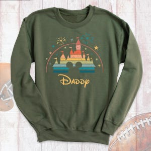May include: Green sweatshirt with a rainbow-colored castle graphic and the word "Daddy" underneath. The castle graphic is a stylized version of the iconic Disney castle.
