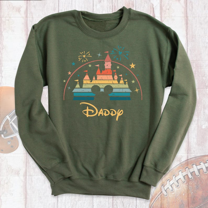 May include: Green sweatshirt with a rainbow-colored castle graphic and the word "Daddy" underneath. The castle graphic is a stylized version of the iconic Disney castle.