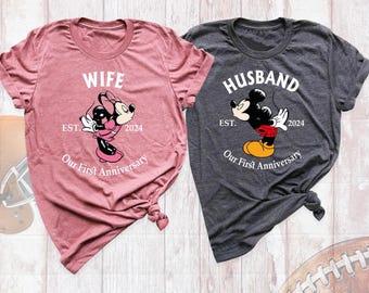 May include: A mauve t-shirt with a graphic of Minnie Mouse and the text WIFE EST. 2024 Our First Anniversary, and a dark gray t-shirt with a graphic of Mickey Mouse and the text HUSBAND EST. 2024 Our First Anniversary.