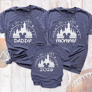 May include: Three dark blue heathered t-shirts featuring a white graphic of a castle silhouette with a Mickey Mouse head shape, fireworks, and stars. One shirt says DADDY, one says mommy, and the smaller shirt displays the year 2026.