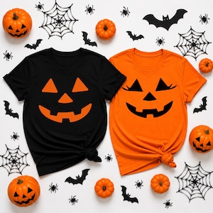 May include: Two short-sleeve t-shirts, one black and one orange, each featuring a large jack-o'-lantern face graphic. The black shirt has an orange pumpkin face, while the orange shirt has a black pumpkin face with eyelashes.