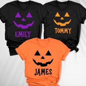 May include: Three short-sleeved t-shirts featuring jack-o'-lantern faces. Two black shirts display purple and orange faces with the names Emily and Tommy. One orange shirt displays a black face with the name James printed below.