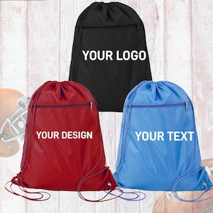 May include: Three drawstring backpacks in black, red, and blue. Each bag features a front zippered pocket and a drawstring closure. The black bag displays the text YOUR LOGO, the red bag displays YOUR DESIGN, and the blue bag displays YOUR TEXT.