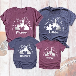 May include: Four short-sleeve t-shirts in maroon and heather navy blue. Each shirt features a white graphic of a castle silhouette with a mouse ear icon, fireworks, and stars. The shirts are labeled with the text Mommy, Daddy, Brother, and Sister.