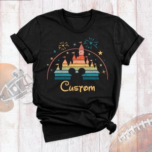 May include: A black short-sleeve t-shirt featuring a graphic of a castle silhouette with a Mickey Mouse head outline, colorful horizontal stripes, fireworks, and stars. The word Custom is printed in a yellow script font below the castle.