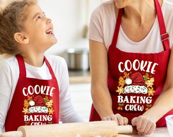 May include: Two red aprons with the text "COOKIE BAKING CREW" and a Santa hat design. A child and an adult are wearing the aprons while baking cookies. A rolling pin and dough are visible.