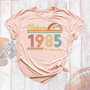 May include: A light pink t-shirt with a vintage-style graphic that says "Vintage 1985 Fort Lauderdale". The graphic features a rainbow with palm trees and is in a retro color palette.