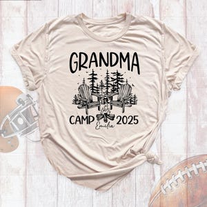 May include: Beige t-shirt with the words "GRANDMA CAMP 2025" in black. The design includes two Adirondack chairs, trees, and a campfire. The name "Emilia" is written below the campfire.