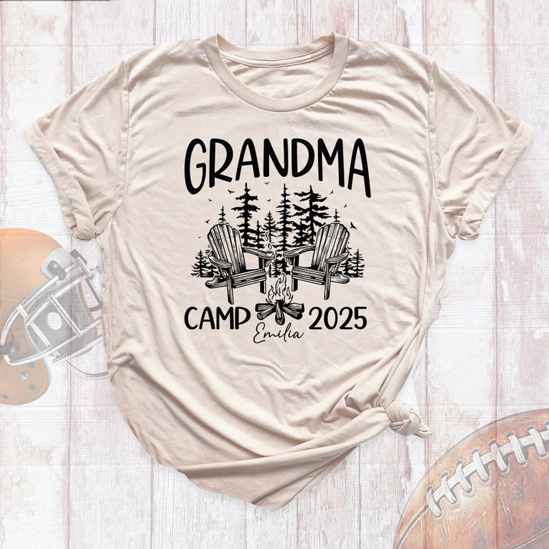 May include: Beige t-shirt with the words "GRANDMA CAMP 2025" in black. The design includes two Adirondack chairs, trees, and a campfire. The name "Emilia" is written below the campfire.