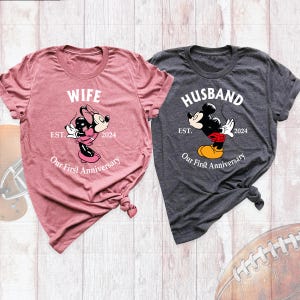 Puede incluir: Dos camisetas, una rosa y otra gris. La camiseta rosa dice "Wife" con una imagen de Minnie Mouse y el texto "EST. 2024 Our First Anniversary". La camiseta gris dice "Husband" con una imagen de Mickey Mouse y el texto "EST. 2024 Our First Anniversary".