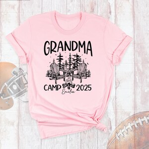May include: A light pink t-shirt with the words "GRANDMA CAMP 2025" in black. The design includes two wooden chairs, a campfire, and trees. The name "Emilia" is printed below the campfire.